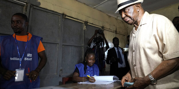 Joseph Boakai - mit Hut - gibt seine Stimme in einem Wahllokal ab. Eine Wahlhelferin sitzt und trägt etwas in ein Heft ein, ein anderer Wahlhelfer beobachtet die Szene stehend. Im Hintergrund Männer, die fotografieren