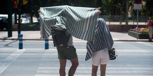 Zwei Menschen stehen an einem Zebrastreifen, sie schützen sich mit Badetüchern vor dre Hitze