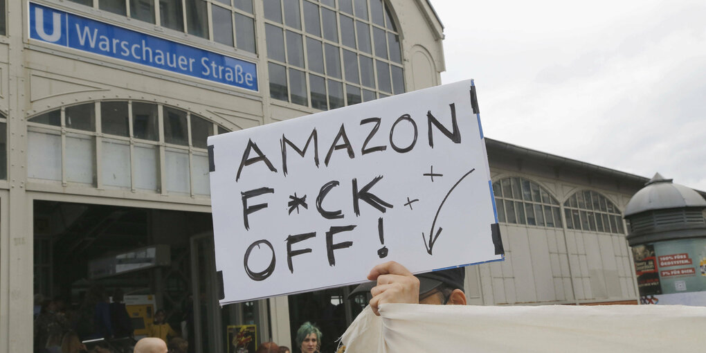 Auf der Demonstration "Ihr habt uns die Stadt gestohlen gebt sie wieder her" vom 9.7.2022 hält ein Mann ein Schild hoch auf dem zu lesen ist "Amazon F*CK OFF!". Im Hintergrund ist das Schild der U-Bahn-Station Warschauer Straße zu sehen.