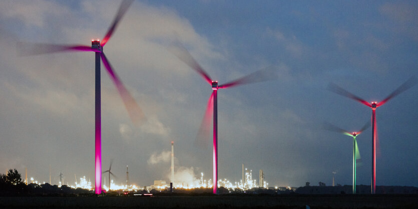 Windräder vor einer beleuchteten Raffinerie.