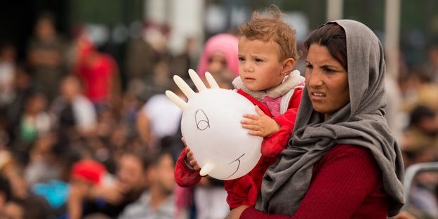 Eine Frau hält ein Kind im Arm. Das Kind hält einen Luftballon mit Gesicht, der aber nur ein aufgeblasener Gummihandschuh ist.