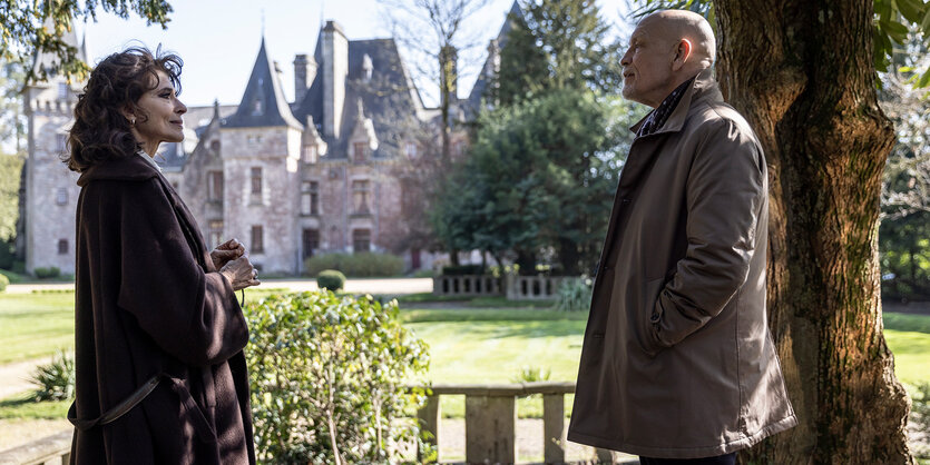 Filmstill aus „Monsieur Blake zu Diensten“: Eine Gutsherrin (Fanny Ardant, links im Bild) und ihr Gast (John Malkovich, rechts im Bild) stehen sich in einem Park gegebüber und unterhalten sich. Im Hintergrund ist ein großes Anwesen zu sehen.