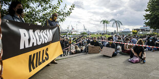 Am Rande einer Versammlung stehen Menschen mit einem Banner, auf dem zu lesen ist "Racism Kills", also "Rassimus tötet"