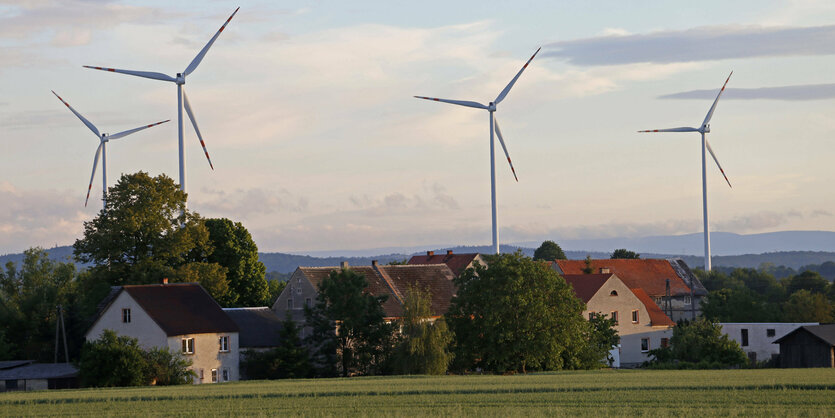 Häuser eines Dorfes, im Hintergrund vier Windkraftanlagen, dahinter weite Waldlandschaft