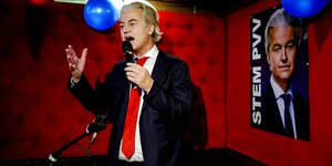 Geert Wilders trägt eine rote Krawatte zu einem blauen Anzug und spricht vor einer roten Wand