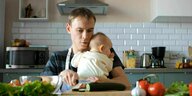 Junger Vater schneidet Gemüse und hält ein Baby in den Händen