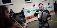 Aktivist:innen spielen vor einem Haus Geige und Akkordeon, im Hintergrund ein Plakat