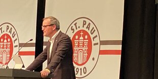 Oke Göttlich steht in einem braunen Anzug an einem Rednerpult, im Hintergrund große Banner mit dem Logo des FC St. Pauli