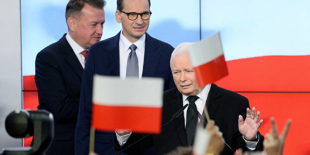 PiS-Chef Jaroslaw Kaczynski winkt auf einer Bühne stehend verhalten ins Publikum. Im Vordergrund leicht unscharf polnische Flaggen, die geschwenkt werden.