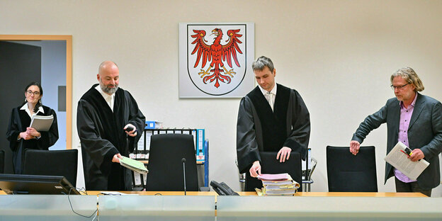 Drei Männer und eine Frau in schwarzer Robe versammeln sich in einem Gerichtsaal