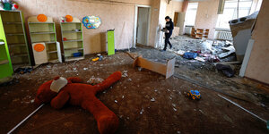 Ein zerstörter Innenraum eines Kindergartens. Auf dem Boden liegt eine große Plüschfigur. Eine Frau räumt Sachen weg