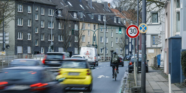 Auf einer Straße mit Tempo-30-Schild fahren Autos an einem Menschen auf dem Fahrrad vorbei. Die Straße säumen Häuserreihen..