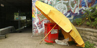 An einem Müllbehälter an einer Unterführung steht ein alter aufgespannter Sonnenschirm.
