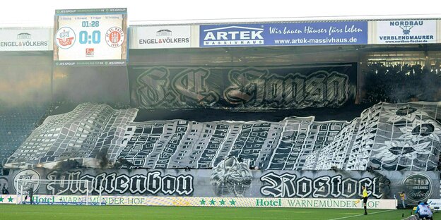 Blick auf die Fankurve eines Fußballstadions, in der ein Banner gehalten wird, auf dem die Plattenbauten von Rostock-Lichtenhagen abgebildet sind