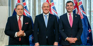 Die neue Regierung in Neuseelan, drei Männer in Anzügen stehen vor grünen Sesseln, hinter ihnen drei Flaggen Neusseelands und in der Mitte lächelt der neue Premierminister Christopher Luxon