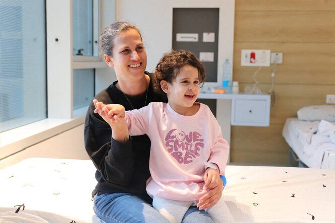 Ein kleines Mädchen in rosa Pullover sitzt auf dem Schoß einer Frau auf einem Krankenhausbett