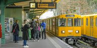Oberirdischer Bahnsteig in Berlin, zwei gelbe U-Bahnen sind zu sehen, vor allem junge Menschen stehen auf dem Bahnsteig und warten auf den einfahrenden Zug