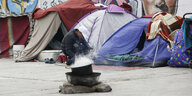 Vor einer bemalten Mauer stehen Zelte, ein Mann hockt vor einem dampfenden Kochtopf