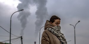 Eine Frau bedeckt ihr Gesicht mit einem Wollschal, im Hintergrund rauchen Schornsteine
