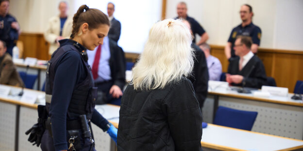 Eine Frau mit weißem langen Haar wird von einer Polizistin in einen Gerichtssaal geführt. Man erkennt sie nicht, nur ihre Haare sind zu sehen.