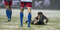 ein Fußballspieler in braunem Dress liegt auf verschneitem Rasen, neben ihm gehen Spieler mit weißen Trikots und roten Hosen