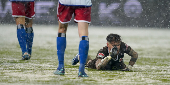 ein Fußballspieler in braunem Dress liegt auf verschneitem Rasen, neben ihm gehen Spieler mit weißen Trikots und roten Hosen