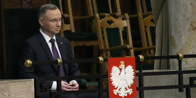 EIn Mann sitzt neben einem Wappen mit Adler auf rotem Grund