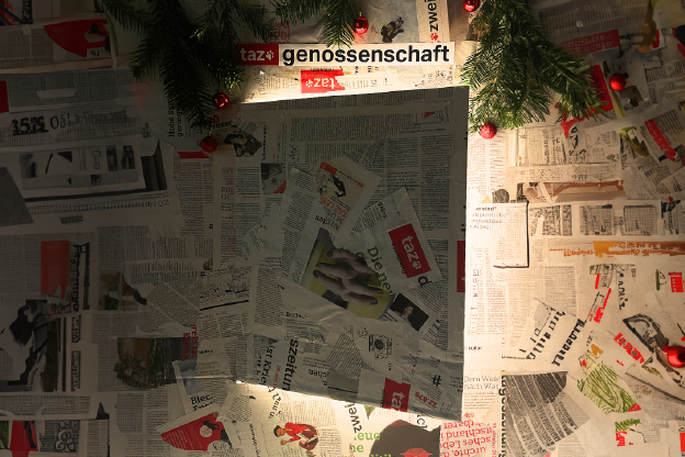 Eine in Zeitungspapier gekleidete Tür geht auf, hinter der Tür leuchtet es. Über die Tür hängt Tannengrün mit Weihnachtskugeln.