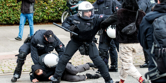 Ein Mensch liegt am Boden, ein Polizist fixiert ihn, ein anderer hat einen Knüppel in der Hand