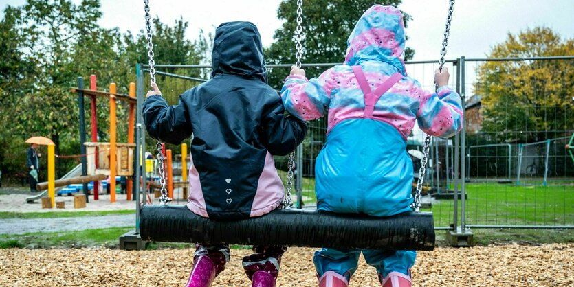 Zwei Kinder sitzen gemeinsam in Regenkleidung auf einer breiten Schaukel