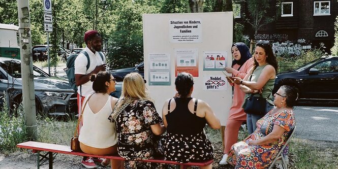 Eine Gruppe Menschen im Hamburger Stadtteil Veddel, die vor einem Aufsteller mit ersten Ergebnissen einer Stadtteilumfrage stehen und sitzen und diskutieren.