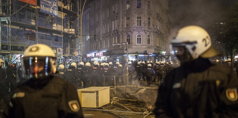 Zwei Polizisten im Vordergrund, noch mehr dahinter auf der Straße in Hamburg. Es ist dunkel, es sind Rauchwolken zu sehen.