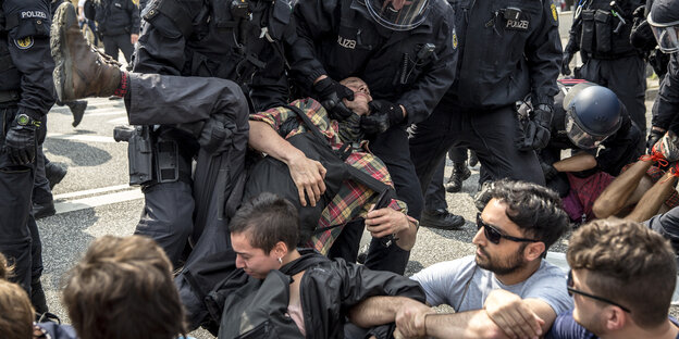 Polizisten halten einen Mann gewaltvoll fest, mehrer Demonstranten sitzen drumherum