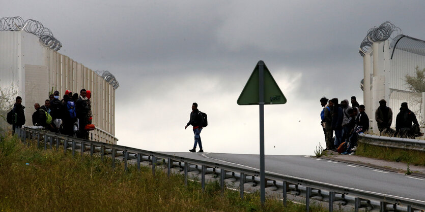 Ein Mensch mit Rucksack geht auf einer Straße zwischen Mauern mit Stacheldraht, an den Seiten stehen viele Menschen