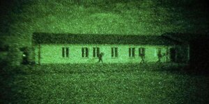 Angehörige des KSK bei einer Nachtübung. Das Bild ist grünlich eingefärbt, wie durch eine Nachtsichtkamera geschossen. Bewaffnete Männer rennen um ein Haus herum.