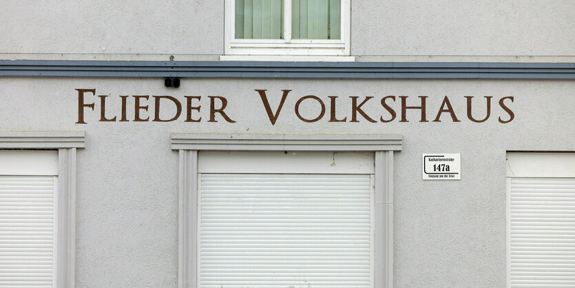 Ein hellgraues Haus mit geschlossenen Rolläden, darüber die Worte: "Flieder Volkshaus"