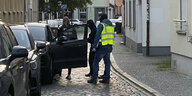 Drei Polizisten, zwei davon vermummt stehen vor einem geparkten Auto mt geöffneter Wagentür