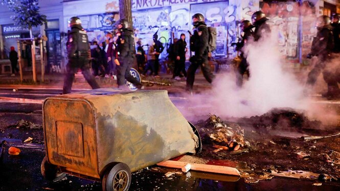 Eine brennende Mülltonne liegt in der Nacht auf ener Straße, im Hinterfund Polizei mt Helmen und junge Menschen mit dunkler Kleidung