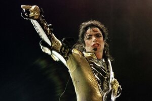 Michael Jackson bei einem Auftritt. Er trägt ein goldenes, eng anliegendes oberteil und hat lange, lockige haare, die nach hinten gebunden sind. Er hat ein Mikrofon an der Wange und streckt den rechten Arm in die Luft.