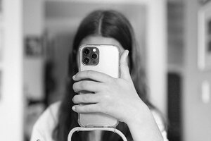 Schwarz-Weiß-Fotografie von einer Frau mit langen, glatten, dunklen Haaren. Man kann ihr Gesicht nicht sehen, weil sie es selbst mit einem Handy verdeckt, das sie vor sich streckt.