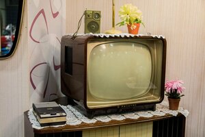 Ein alter Röhrenfernseher mit brauner, holzartiger umkleidung an den Seiten, der auf einem Tischchen mit Tischdecke steht. auf dem Fernseher Mumpitz und ein Radio. Alles wirkt sehr aus der Zeit gefallen.