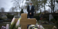 Ein Holzkreuz auf einem Grab, im Hintergrund steht ein Mann