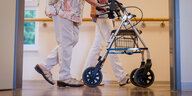 Eine Pflegefachkraft begleitet eine ältere Person, die einen Rollator vor sich herschiebt, durch die Gänge eines Pflegeheimes.
