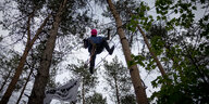 Ein Mann mit rosaner Gesichtsmaske geht auf einem Seil, welches im Wald an den Bäumen befestigt ist
