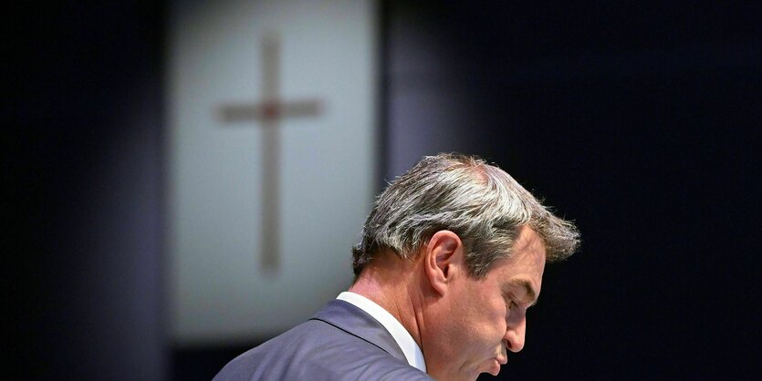 Markus Söder vor einem Holzkreuz, das an einer Wand hängt