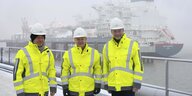 Robert Habeck, Olaf Scholz und Christian Lindner stehen in neongelben Jacken und weißen Helmen vor einem Schiff