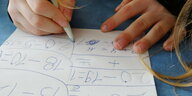 Die Hände eines Kindes halten einen Kugelschreiber über ein Blatt Papier, auf dem Rechenaufgaben geschrieben stehen