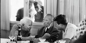 Ben Gurin und Adenauer sitzen an einem Tisch, daneben ein weiterer Mann, Soldaten schauen durch ein Fenster im Hitnergrund