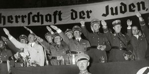 Führende Nationalsozialisten stehen in Uniform auf einer Bühne und zeigen den Hitlergruss - hinter ihnen ein Band: Herrschaft der Juden