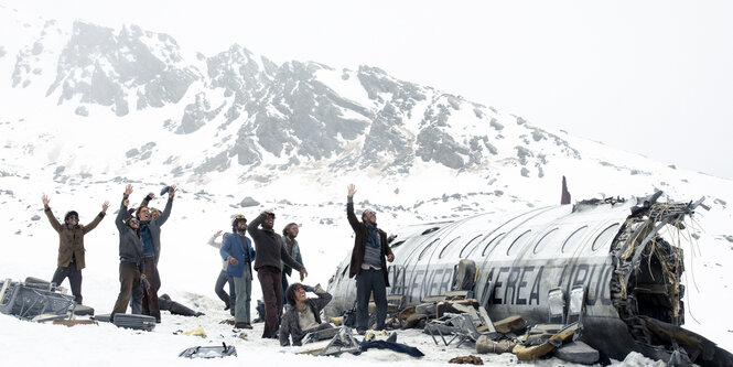 Ein Flugzeugwrack, daneben Überlebende Passagiere, im Hintergrund hohe Berge. Szene aus "Die Schneegesellschaft"